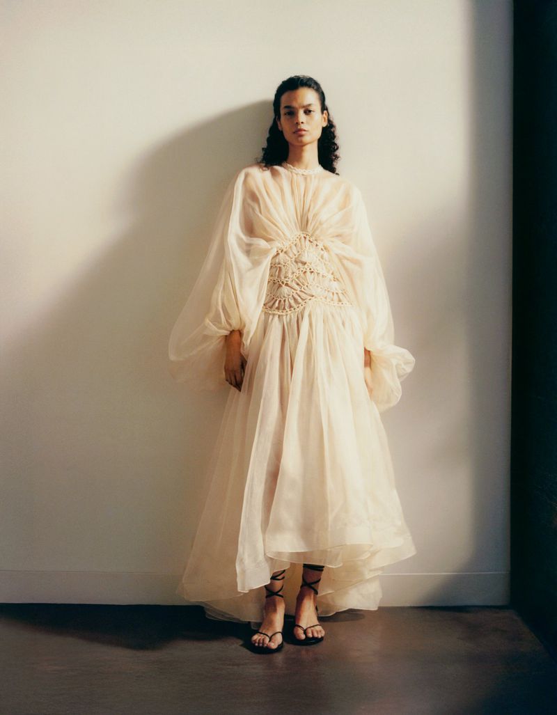 Fashion shot of a woman wearing a long, sheer, ivory dress with a crocheted bodice, standing against a neutral-tone wall in soft lighting.