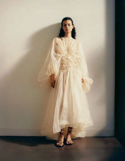 Fashion shot of a woman wearing a long, sheer, ivory dress with a crocheted bodice, standing against a neutral-tone wall in soft lighting.