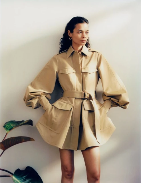 A slender adult female poses in a belted tan jacket-dress with balloon sleeves. The shot is set against a neutral wall with a leafy plant.