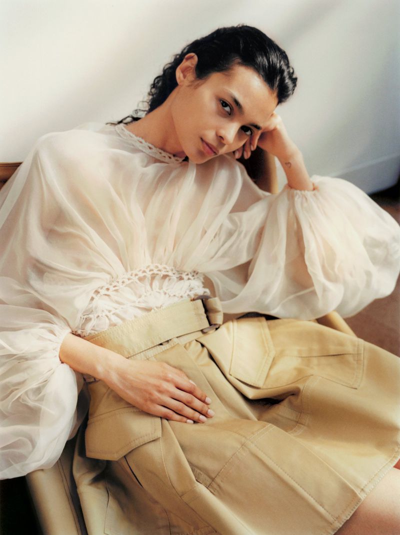 Person with long dark hair poses indoors, wearing a sheer blouse with a crochet neckline and a tan skirt in soft, diffused light.