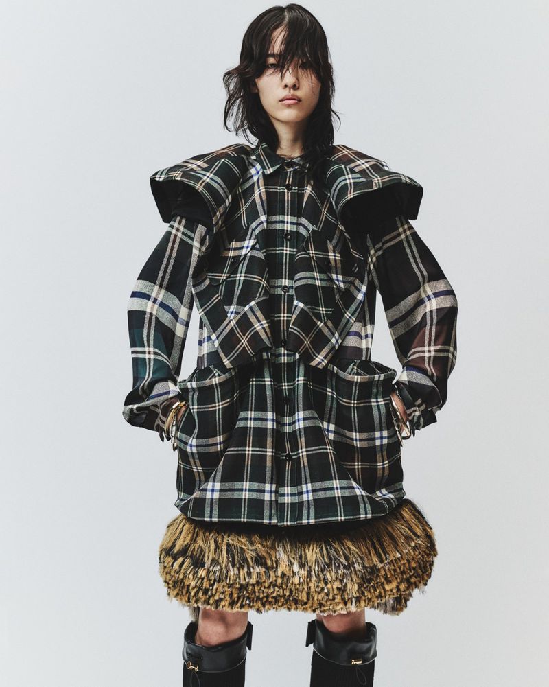 Model wearing a plaid patterned structured jacket and dark knee-high boots against a plain light grey background