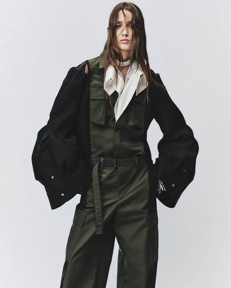 Model poses in an olive green utility jumpsuit with black jacket and white neck scarf against a plain studio background.