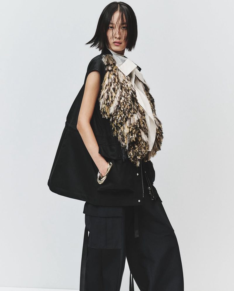Asian woman in black vest with faux fur collar and wide-leg pants, standing in a neutral studio, in a fashion editorial full shot.