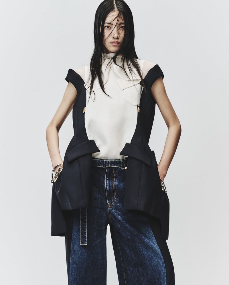 Fashion editorial shot of a slim Asian woman wearing dark-wash denim, a white top, and a long black vest posing against a plain backdrop.