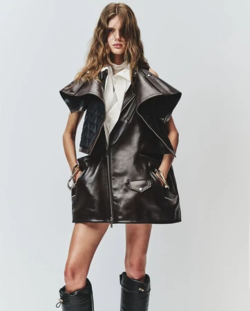 Fashion shot of a young adult female model with long, wavy auburn hair posing in a dark brown leather dress and black leather boots.