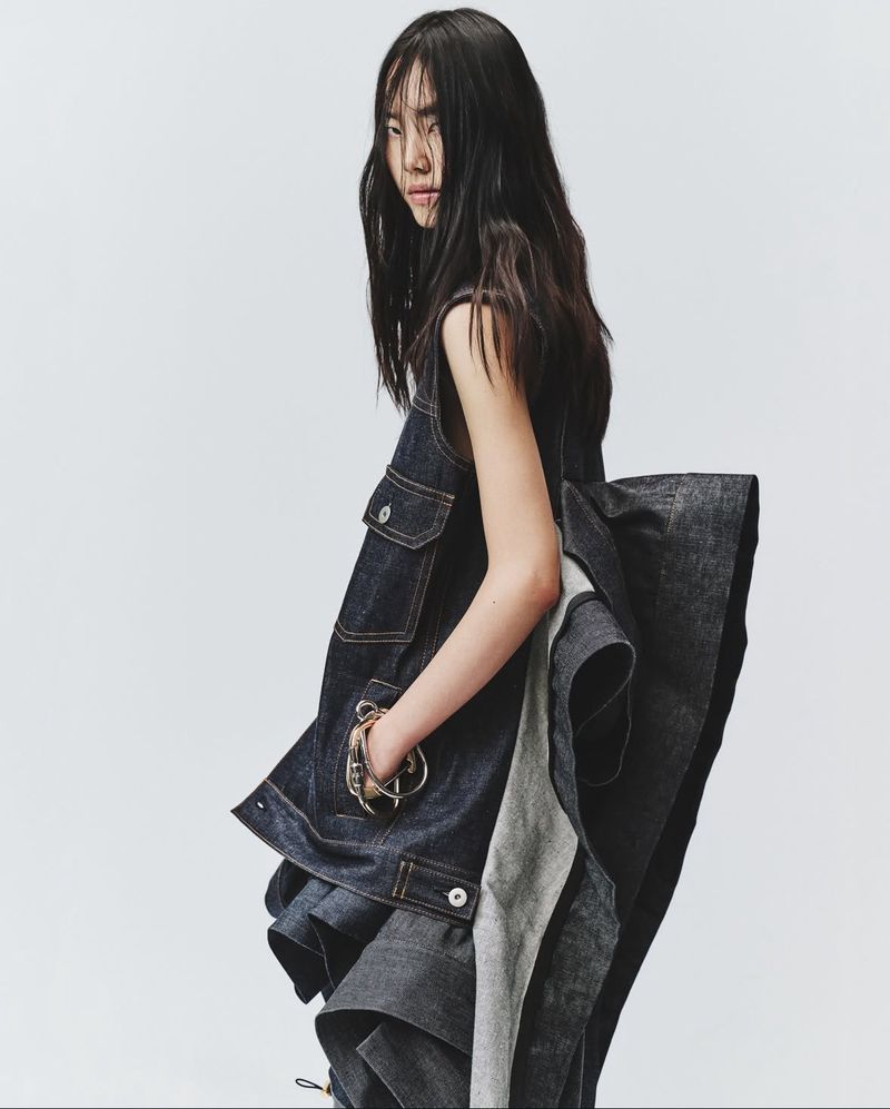 A young Asian woman poses in a sleeveless, deconstructed dark denim dress with long, dark hair against a neutral backdrop, studio lighting.