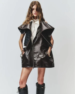 Model wearing a black leather sleeveless jacket over a white shirt and dark boots against a white backdrop.