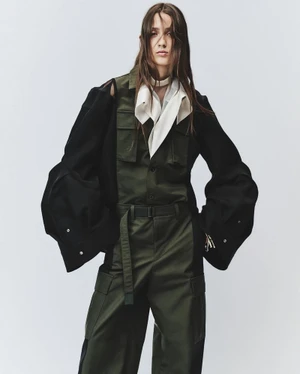 Model in olive utility jumpsuit and black jacket against a plain grey backdrop for Sacai spring summer 2025