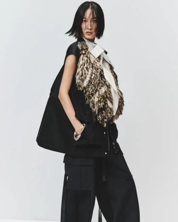 Model in black vest with textured fringe and dark trousers standing against a plain white background for Sacai