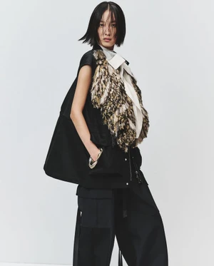 Model in black vest with textured fringe and dark trousers standing against a plain white background for Sacai