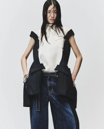 Model wearing a white blouse, black tailored vest, and dark denim jeans against a plain white background.