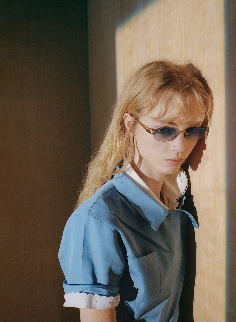 Young woman in a blue top and glasses poses in front of a wall with natural light creating shadows, fashion portrait, studio lighting.