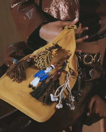 Close-up of tan Miu Miu bag with tassels and charms, held by person in brown outfit. Fashion accessory advertisement, luxury brand.