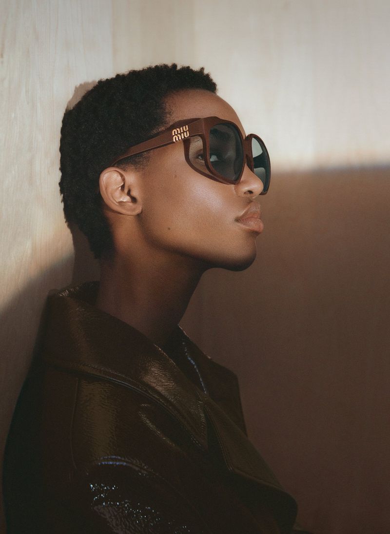 Profile shot of an African-American woman wearing oversized Miu Miu sunglasses and a shiny dark jacket in a softly lit setting.
