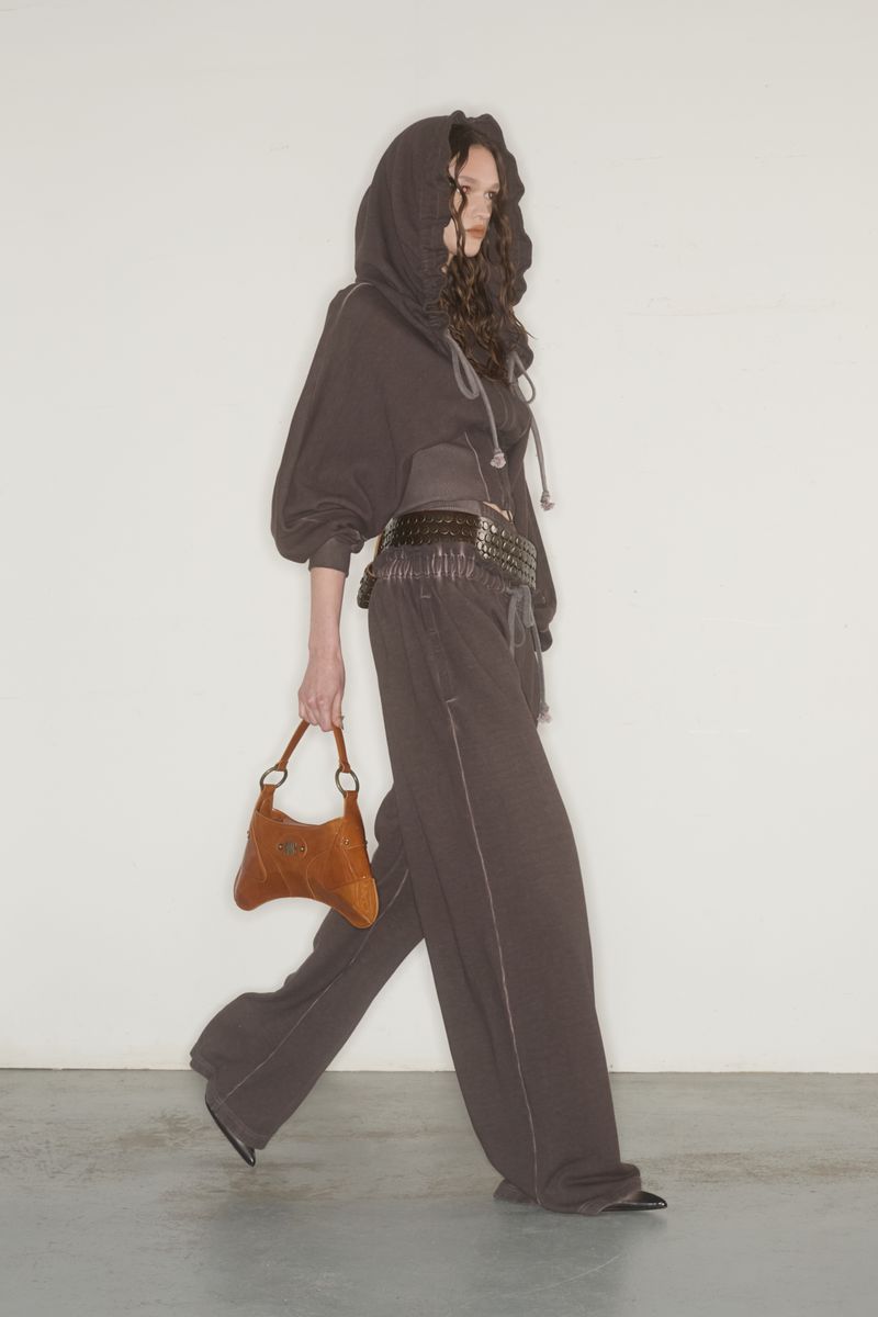 A person walks in a photo studio wearing a hooded top, wide-leg pants, and a brown purse, against a white background.