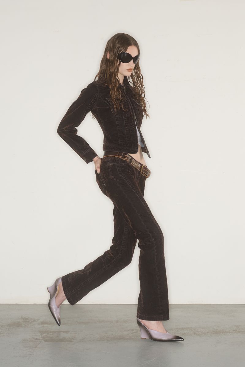 A slender young woman walks in a studio wearing brown corduroy suit with a fitted jacket, matching pants, sunglasses and translucent heels.