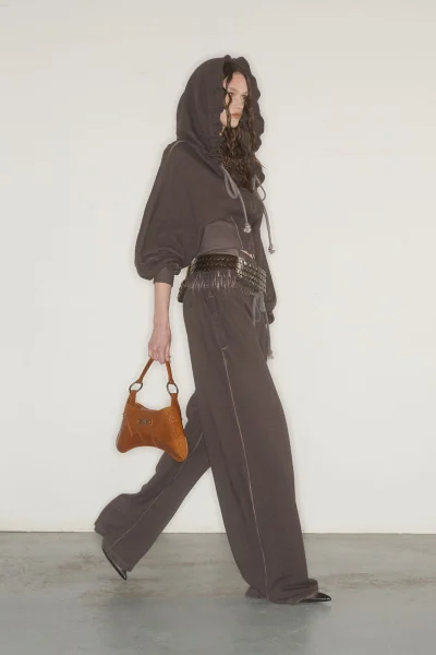 Model wearing a dark grey hooded sweatshirt and wide-leg trousers holding a brown leather handbag against a plain background