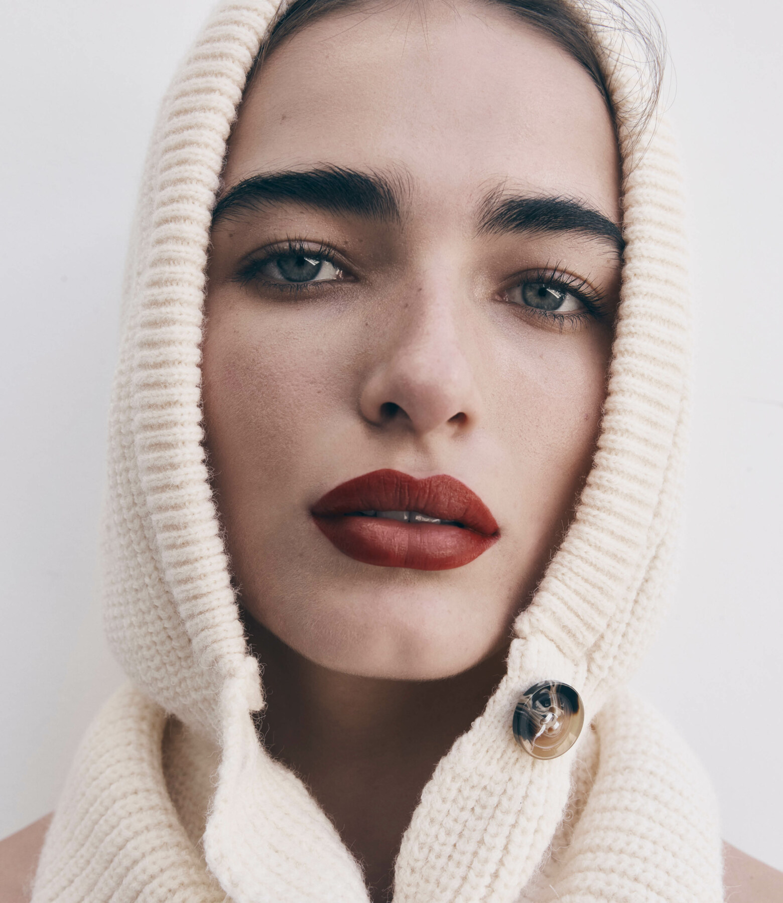 Close-up portrait of a woman wearing a knitted cream-colored hooded scarf, featuring her face with red lipstick against a white background.