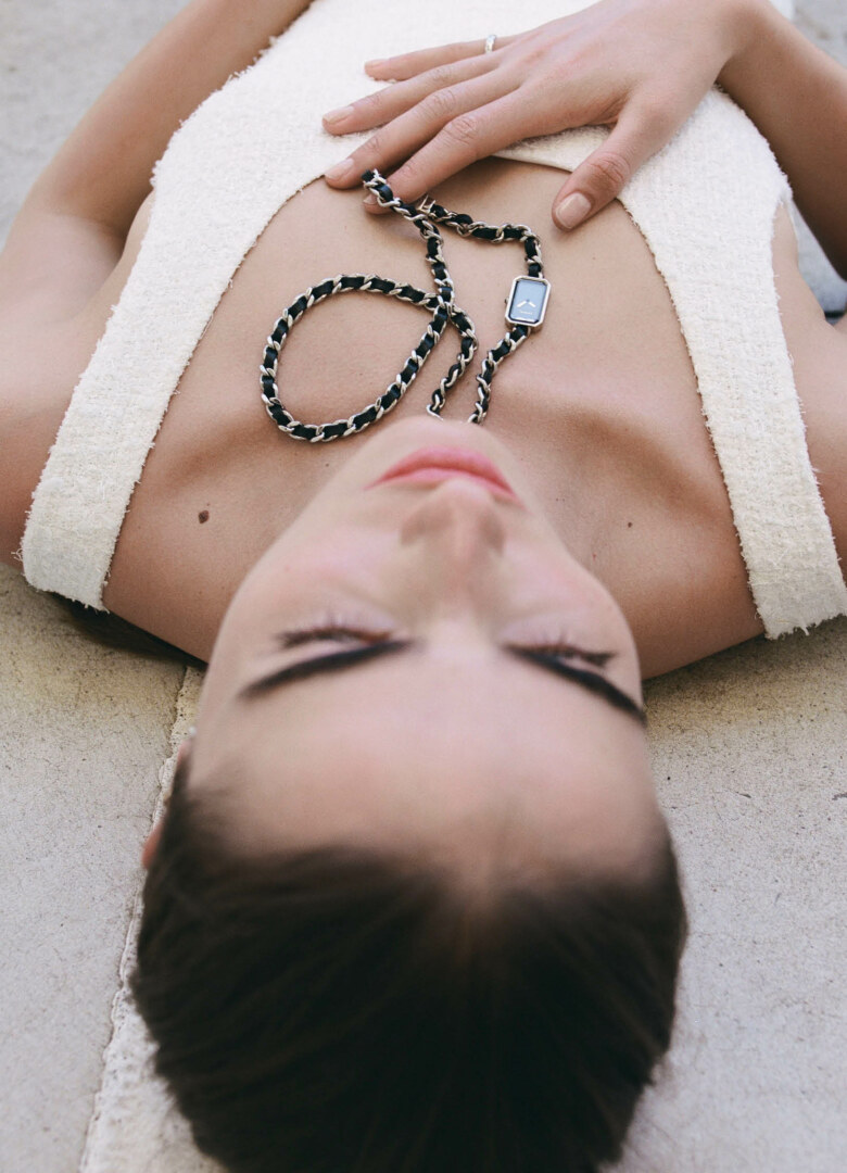 Woman poses in a tank top with a watch necklace, lying on a neutral surface in a top-down view.  Fashion, jewellery, close-up