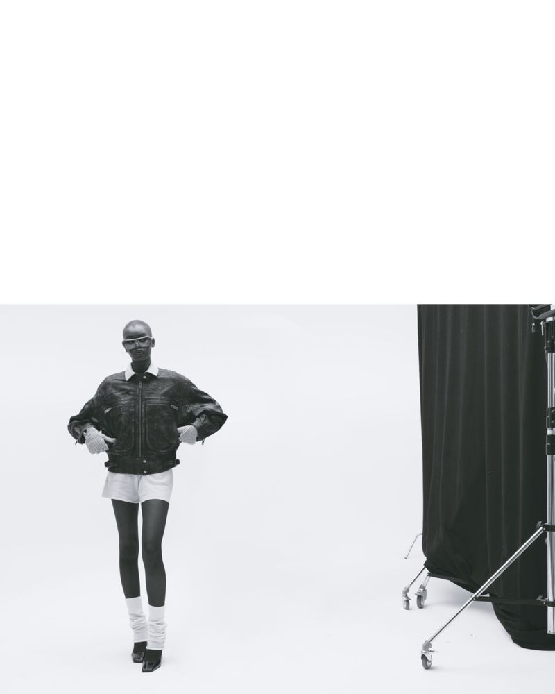 Fashionable person with shaved head in a denim jacket, shorts, and leg warmers posing in a photo studio with a neutral backdrop.