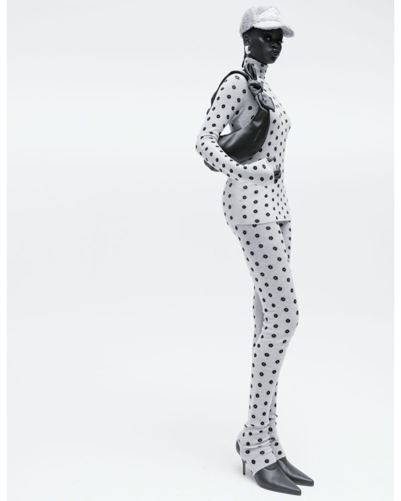 Studio shot of a black female fashion model wearing a polka dot long sleeve shirt, leggings, baseball cap and carrying a purse.