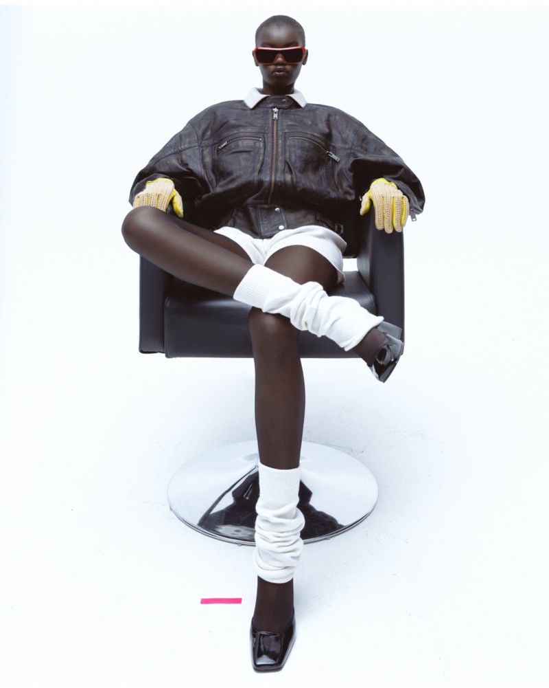 Fashion photo of a Black female model sitting in a black chair wearing sunglasses, jacket, shorts, leg warmers, gloves, and high heels.