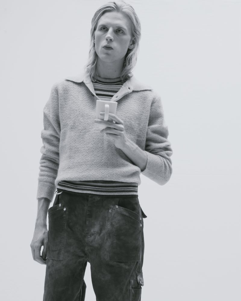 Monochromatic shot of a fair-skinned adult male holding a white mug, wearing a grey sweater and textured pants against a bright backdrop.
