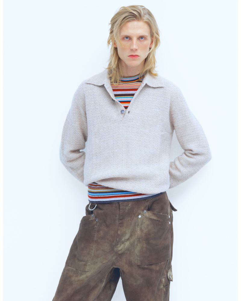 A blond adult male wearing a tan sweater and brown pants poses indoors against a white background, creating a minimalist fashion shot.