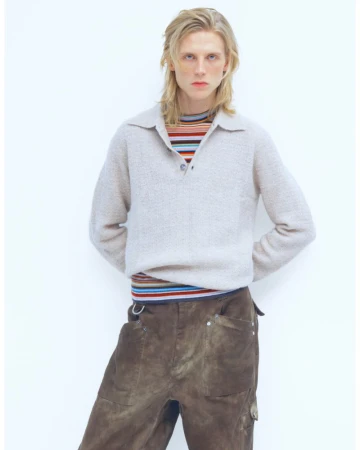 A blond adult male wearing a tan sweater and brown pants poses indoors against a white background, creating a minimalist fashion shot.