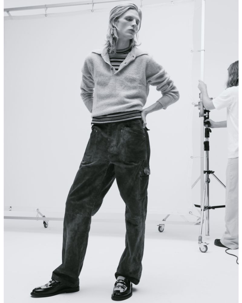 A slender male model with blond hair poses in a photo studio wearing a striped shirt, grey collared sweater, and loose-fitting dark pants.