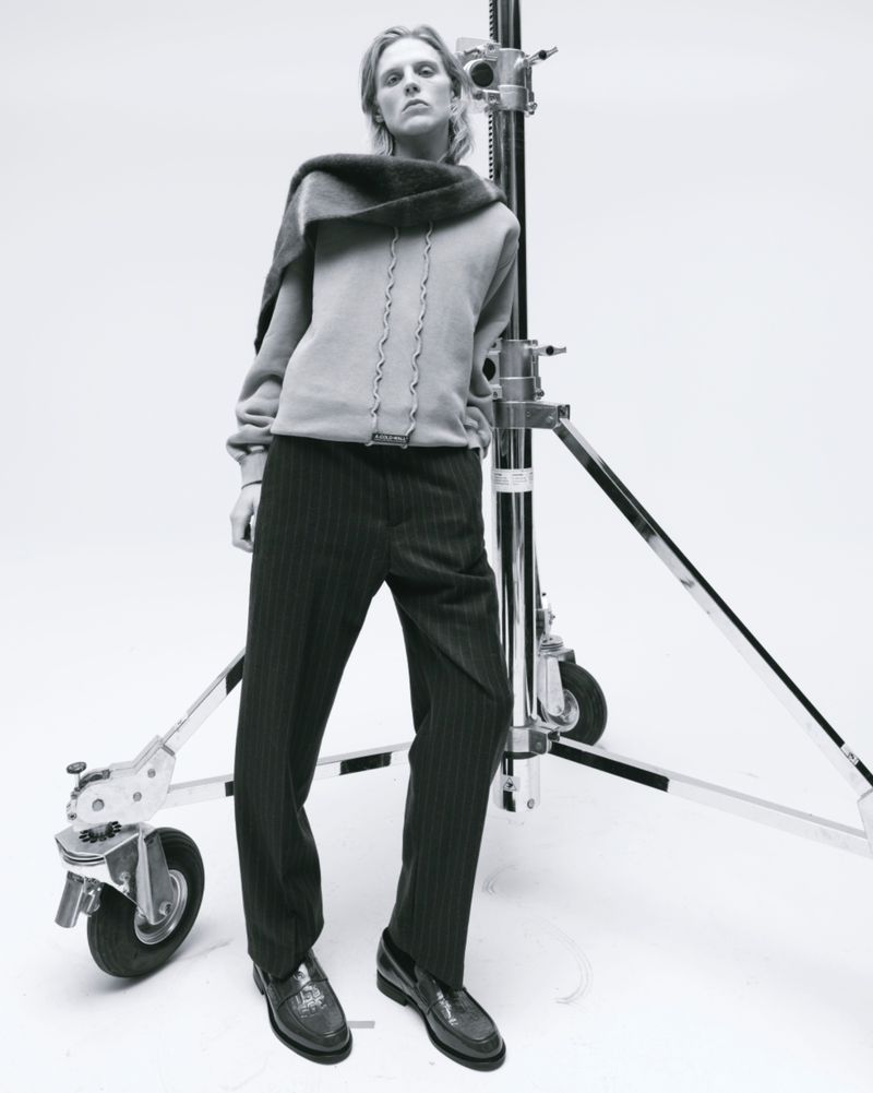Indoor fashion shot of a blonde adult wearing a grey hoodie, pinstripe trousers, and loafers, posed in front of a film light stand.