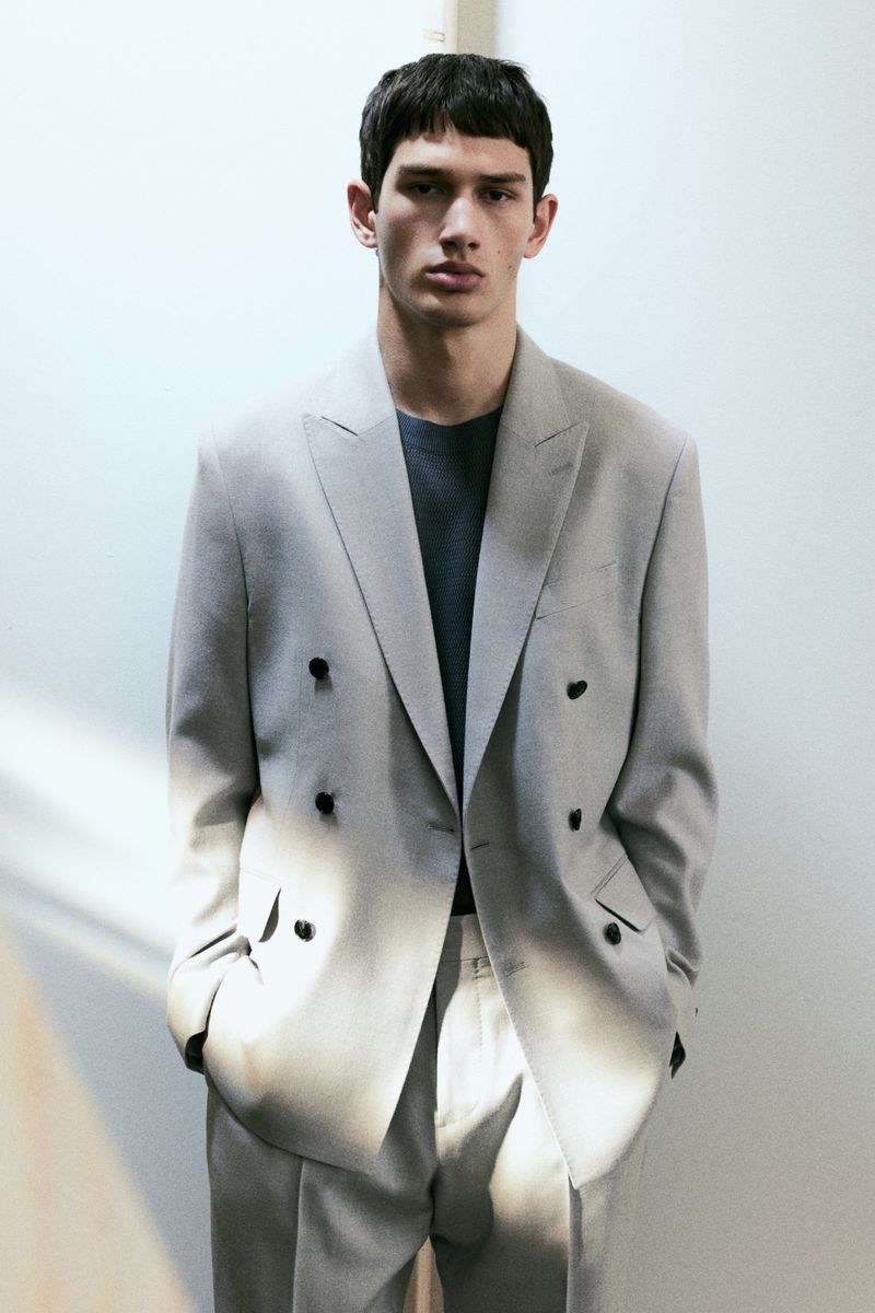 Portrait of a young adult male wearing a gray double-breasted suit, standing indoors against a white wall with natural light.