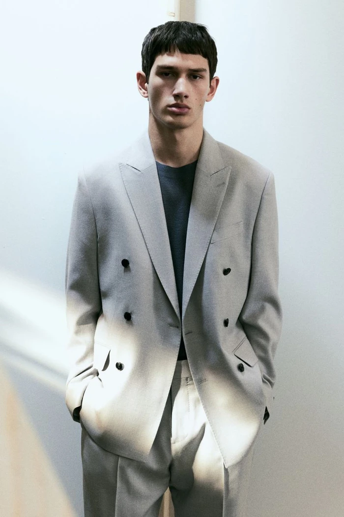Model wearing a grey double-breasted suit jacket over a dark t-shirt against a plain light wall background.
