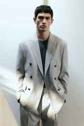 Model wearing a grey double-breasted suit jacket over a dark t-shirt against a plain light wall background.
