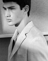 Close-up portrait of a male model wearing a tailored suit jacket against a textured wall in a black and white photograph.