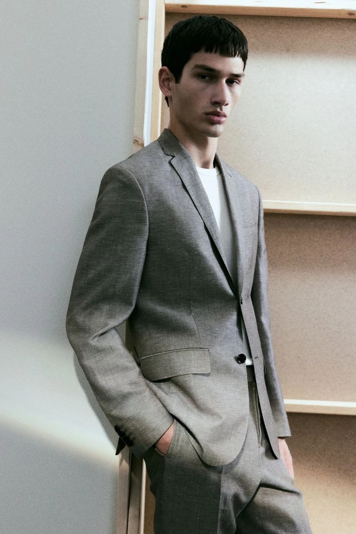 A male model wearing a grey suit jacket over a white t-shirt, posed indoors against a light-coloured wooden frame background.