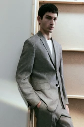 A male model wearing a grey suit jacket over a white t-shirt, posed indoors against a light-coloured wooden frame background.