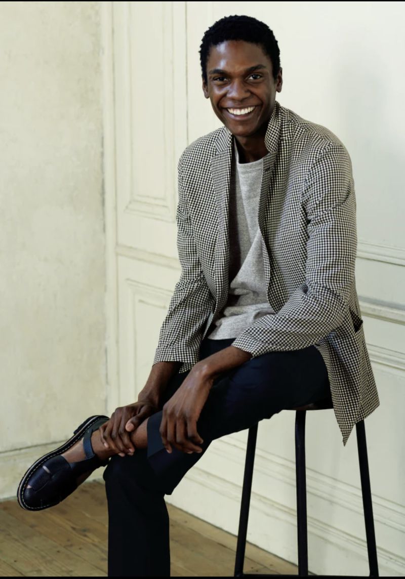 Smiling African-American man in blazer sitting on stool in a studio. He is wearing a grey t-shirt and black pants, fashion portrait.
