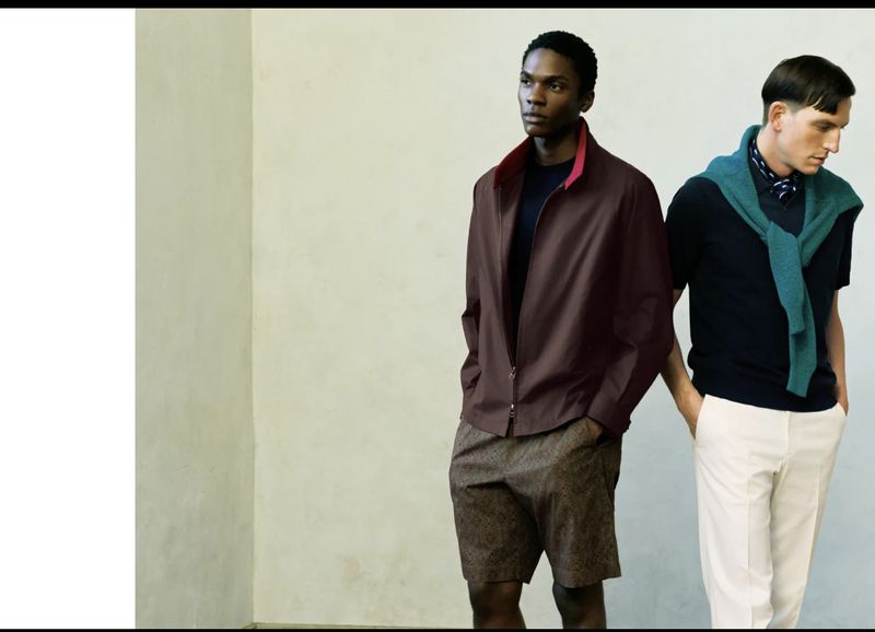 Two male fashion models pose indoors against a neutral wall, showcasing a brown jacket, shorts, navy polo, white pants, and a draped sweater.