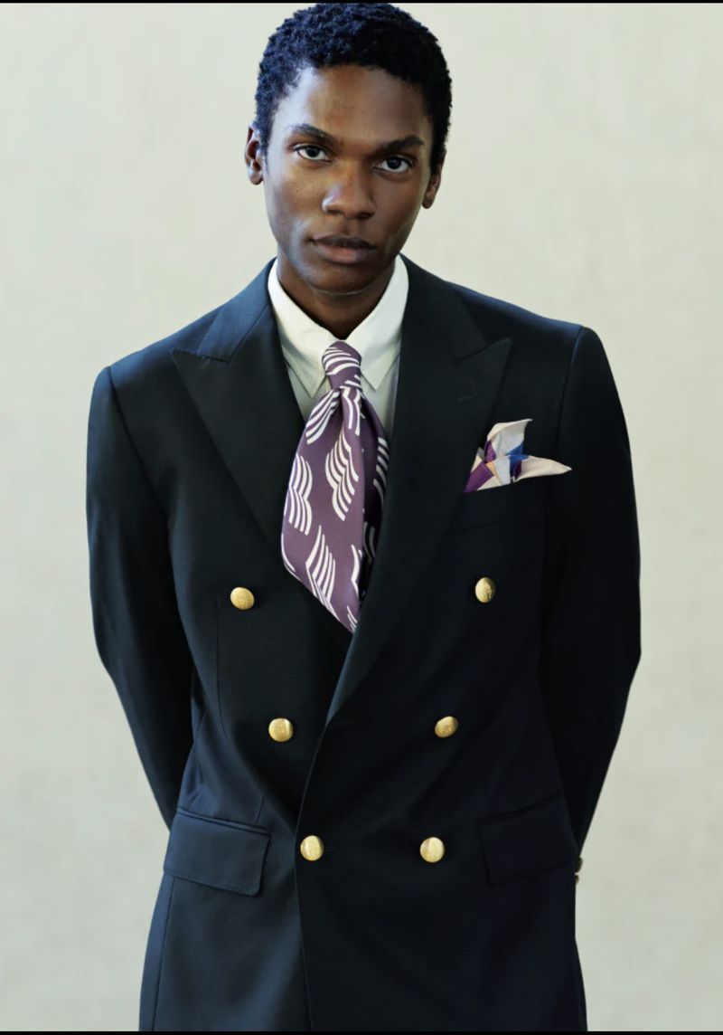 Elegant adult Black male in double-breasted navy suit, gold buttons, patterned tie and pocket square, eye-level indoor studio portrait.