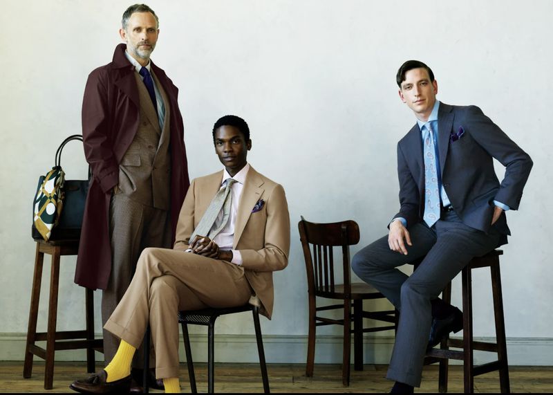 Three male models in tailored suits posing together on wooden stools against a pale neutral wall background.
