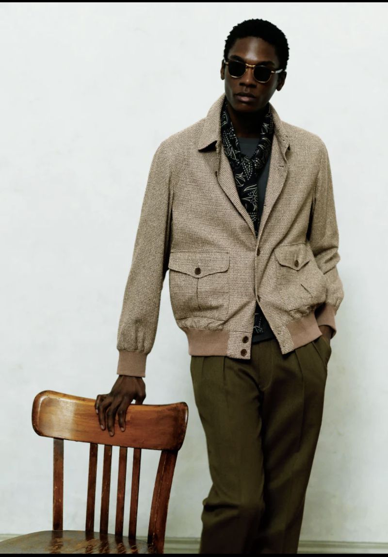 Man in sunglasses poses with a brown jacket, patterned scarf, olive pants, and a wooden chair against a white background.