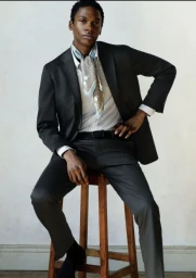 A man wearing a dark suit jacket, white shirt, and patterned neck scarf seated on a wooden stool against a pale wall.