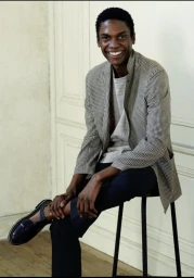 Black man smiling while seated on a stool wearing a patterned blazer and dark trousers