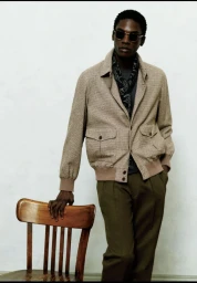 Model wearing a tan jacket, dark sunglasses, and patterned scarf standing with hand resting on a wooden chair.