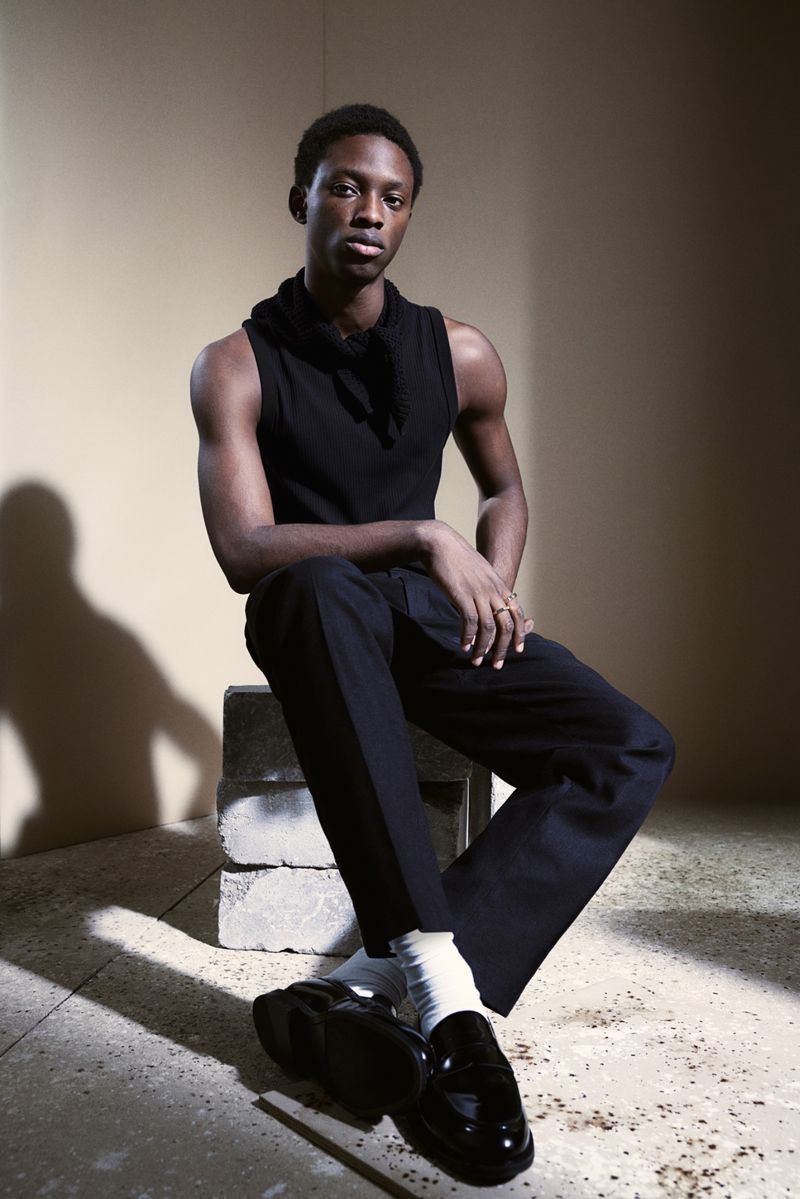 A young black man in a sleeveless top poses seated on concrete blocks indoors with monochrome style against a beige wall.