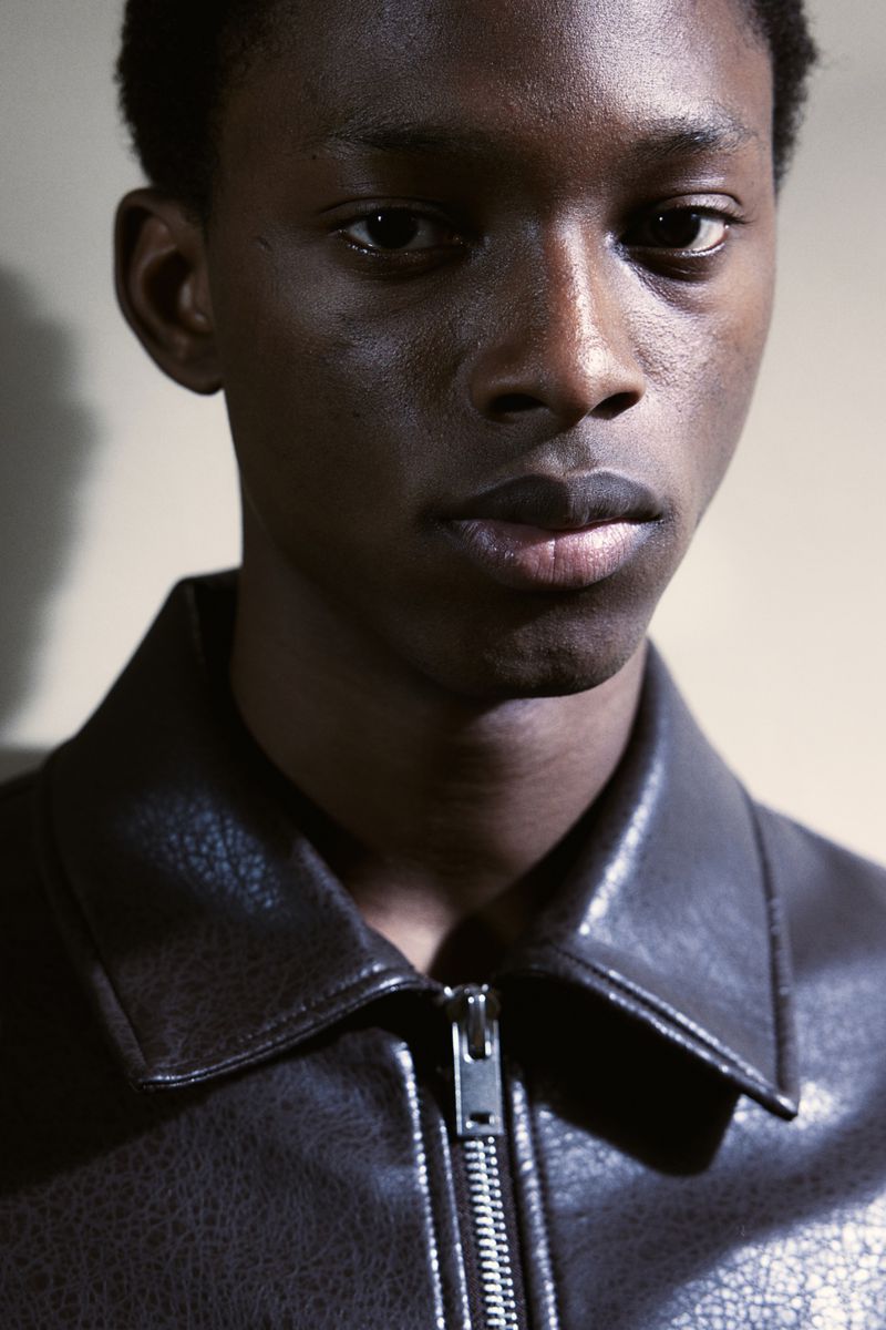 Close up portrait of a Black man wearing a dark textured zip up jacket against a plain wall.