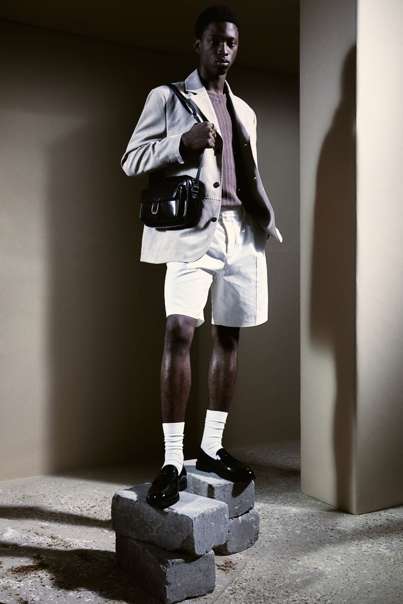 Male fashion model poses in a beige blazer, white shorts, socks and loafers, with a shoulder bag, standing on stacked concrete blocks.