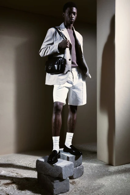 Male fashion model poses in a beige blazer, white shorts, socks and loafers, with a shoulder bag, standing on stacked concrete blocks.