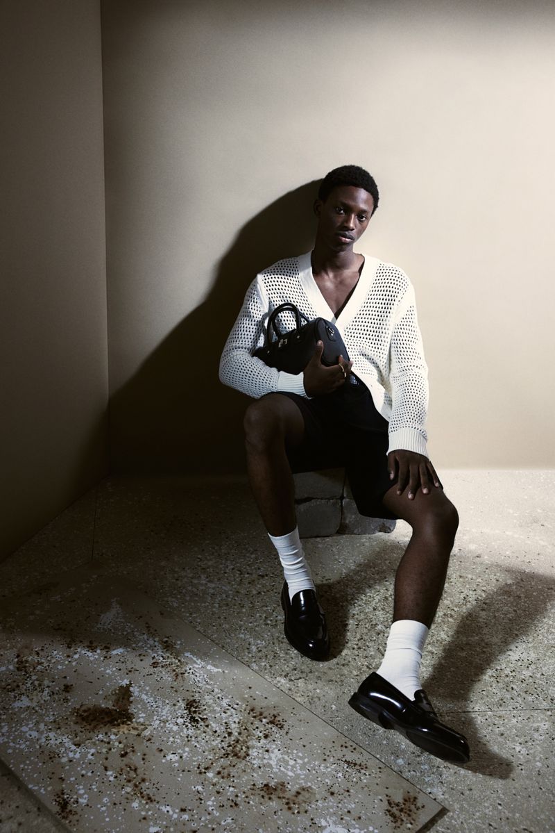 Man in knit cardigan sitting indoors holding dark purse, wearing black shorts and white socks with loafers in soft studio lighting.
